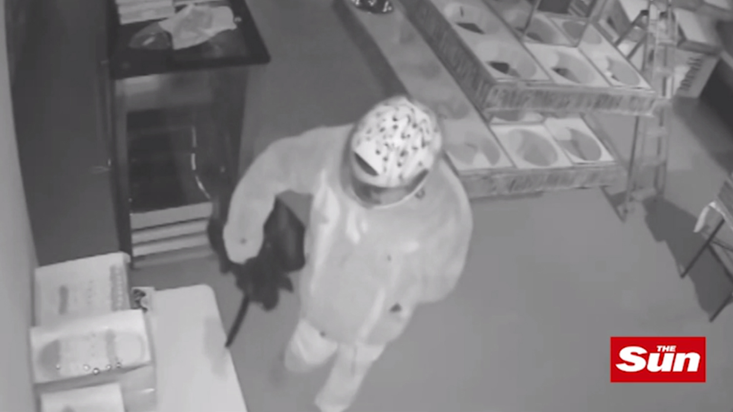 The masked man clamly paces around the kitchen dousing it with a flammable liquid from a black container