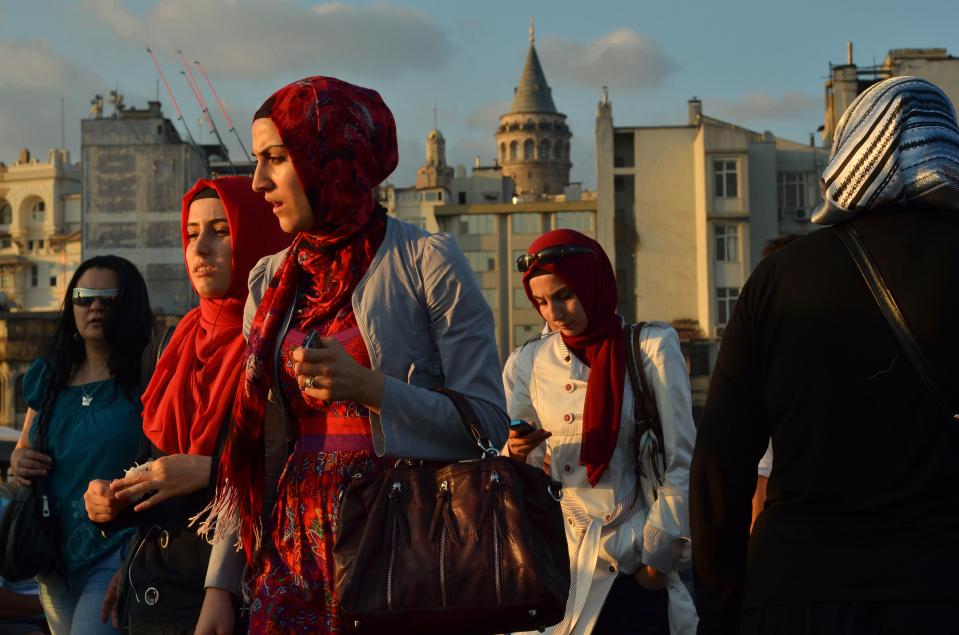 Several Turkish women wearing Turkish headscarves and one woman without head covering walk on Galata bridge at sunset. Galata to