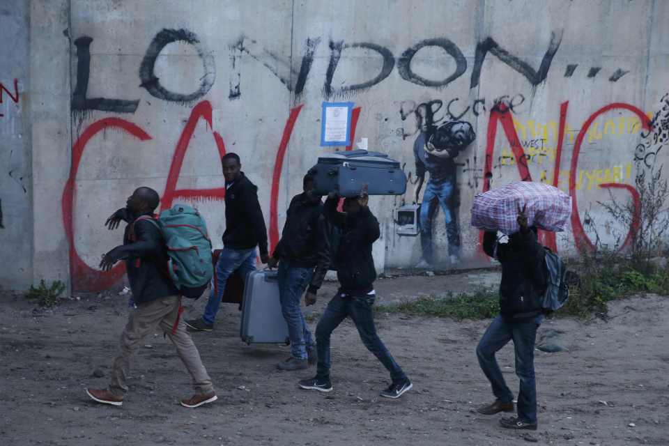 Migrants carry their belongings past a graffiti message reading 'London Calling'