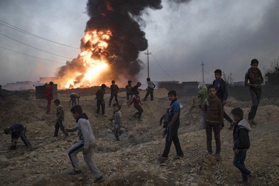 Children play while behind them the oil field in Qayara, south of Mosul, burns
