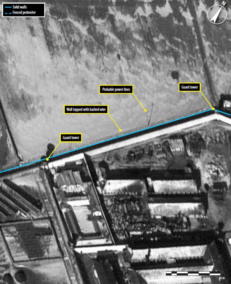 The northwest wall of the light industrial and prisoner housing area, a guard tower, probable power lines and a wall topped with barbed wire 