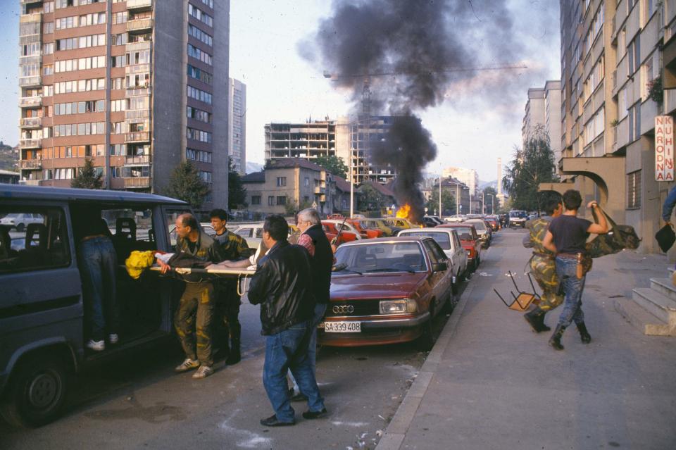 A car burns in the background as a wounded young man is loaded into a car in Sarajevo in 1992