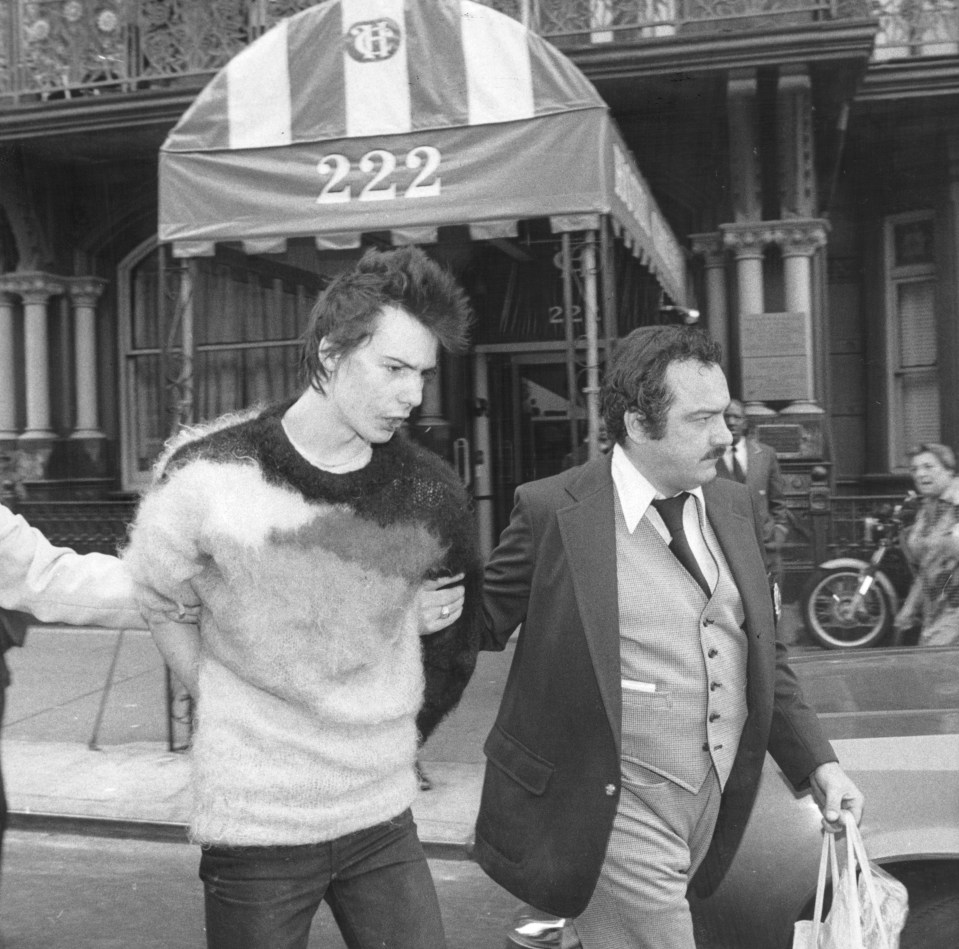 a man in a sweater is being escorted by a man in a suit in front of a building with the number 222 on it