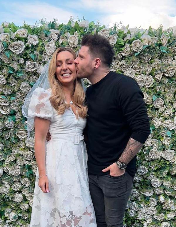 a man kisses a woman on the cheek in front of a wall of flowers
