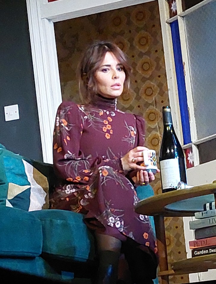 Cheryl in a floral dress holding a mug, sitting on a teal couch.