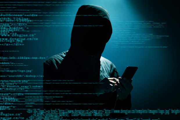 A hooded figure using a smartphone in front of a background of computer code.