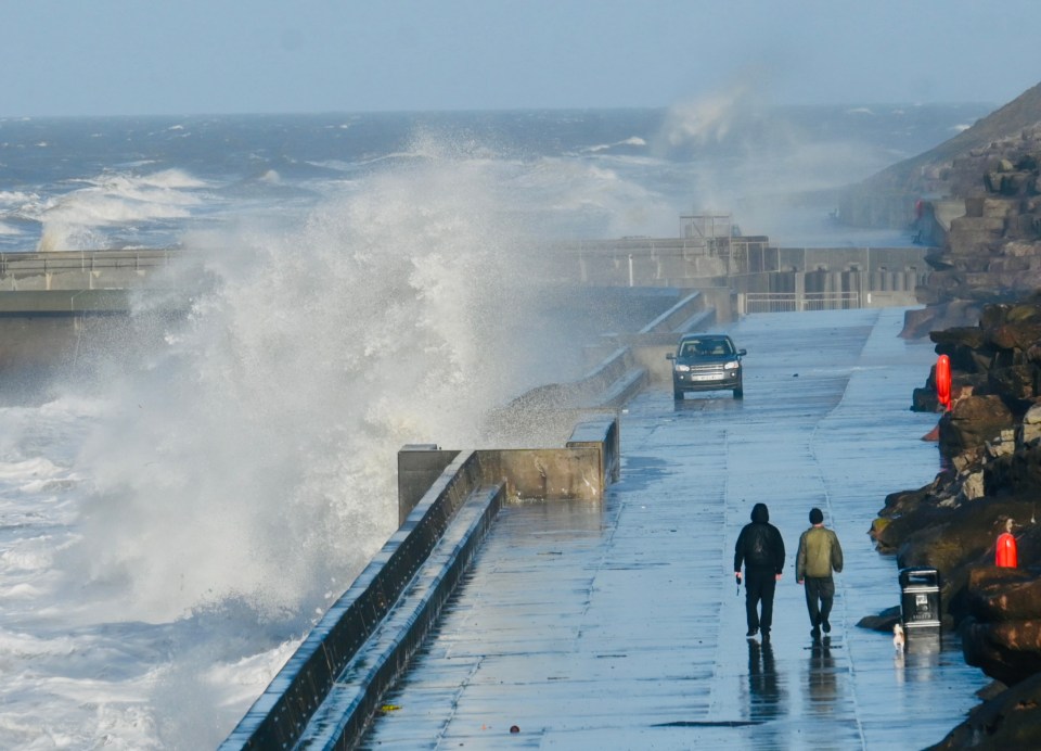 Stormy sea waves crashing against a seawall; two people walk on the wet promenade with a small dog.