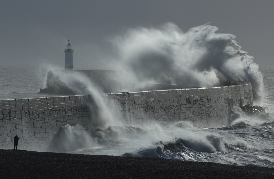 Large waves crashing against a seawall and harbor with a lighthouse in the background.