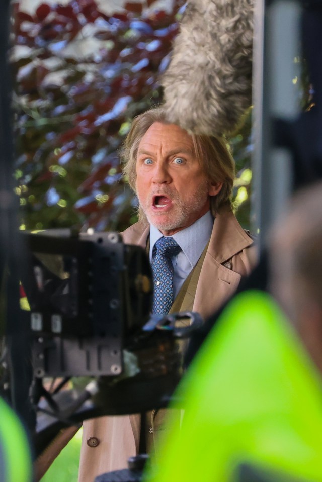 a man in a trench coat and tie stands in front of a camera with his mouth open