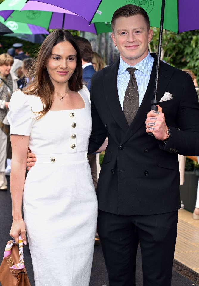 a man in a suit and a woman in a white dress pose under an umbrella that says wimbledon