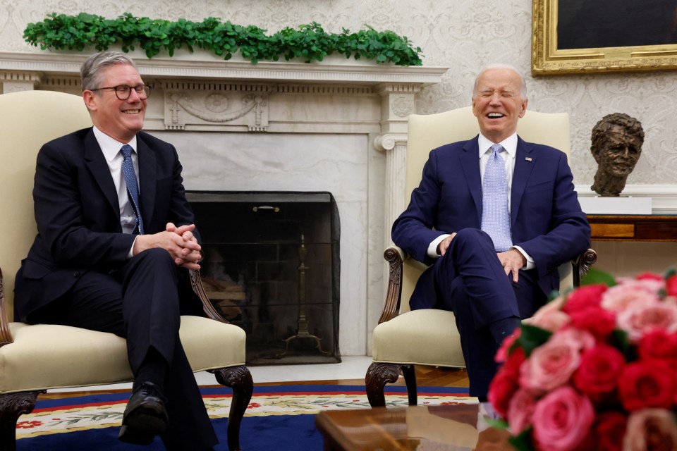 two men in suits sit in chairs in front of a fireplace
