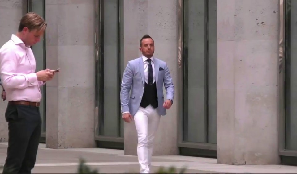 a man in a blue jacket and white pants walks while another man looks at his phone