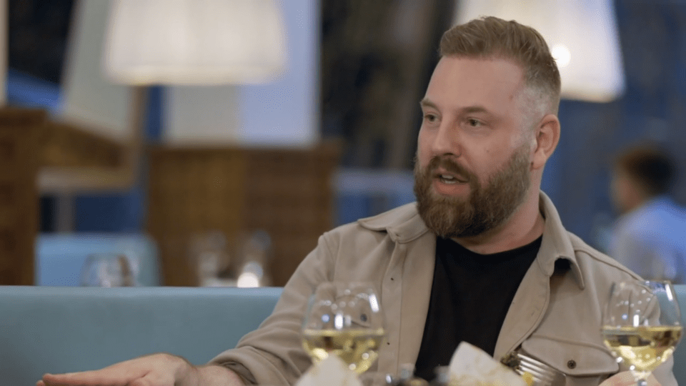a man with a beard sits at a table with two glasses of wine