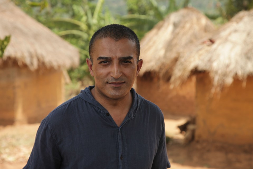 a man in a blue shirt stands in front of a thatched hut