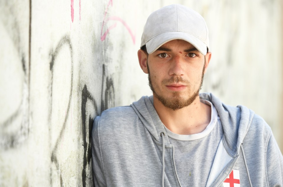 a man leaning against a wall wearing a hat and a hoodie with a cross on it