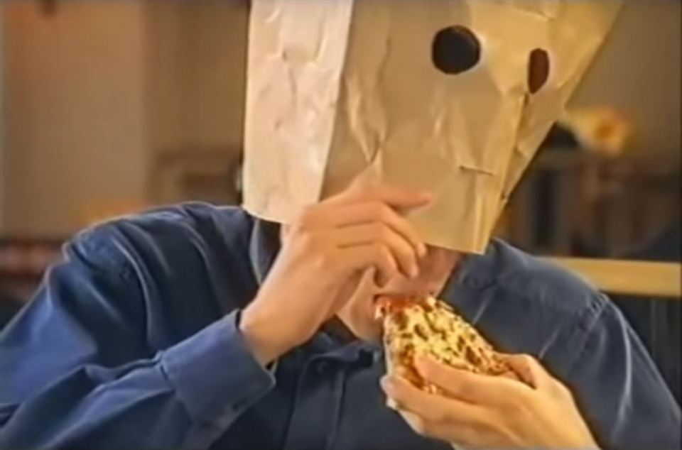 a person wearing a paper bag on their head eating a slice of pizza