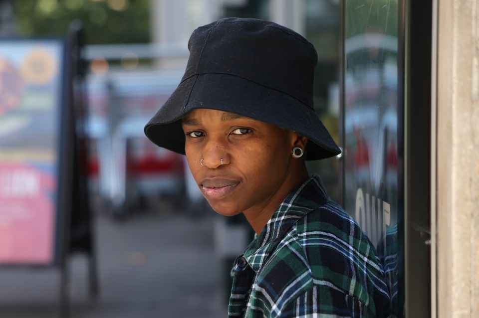 a woman wearing a plaid shirt and a black hat leans against a wall