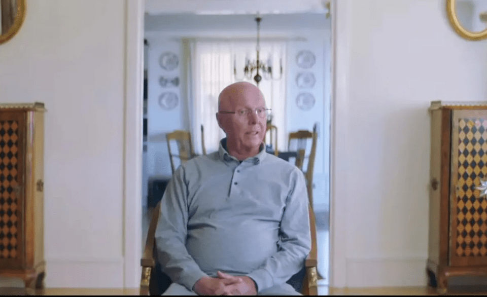 a bald man wearing glasses sits in a chair in a room