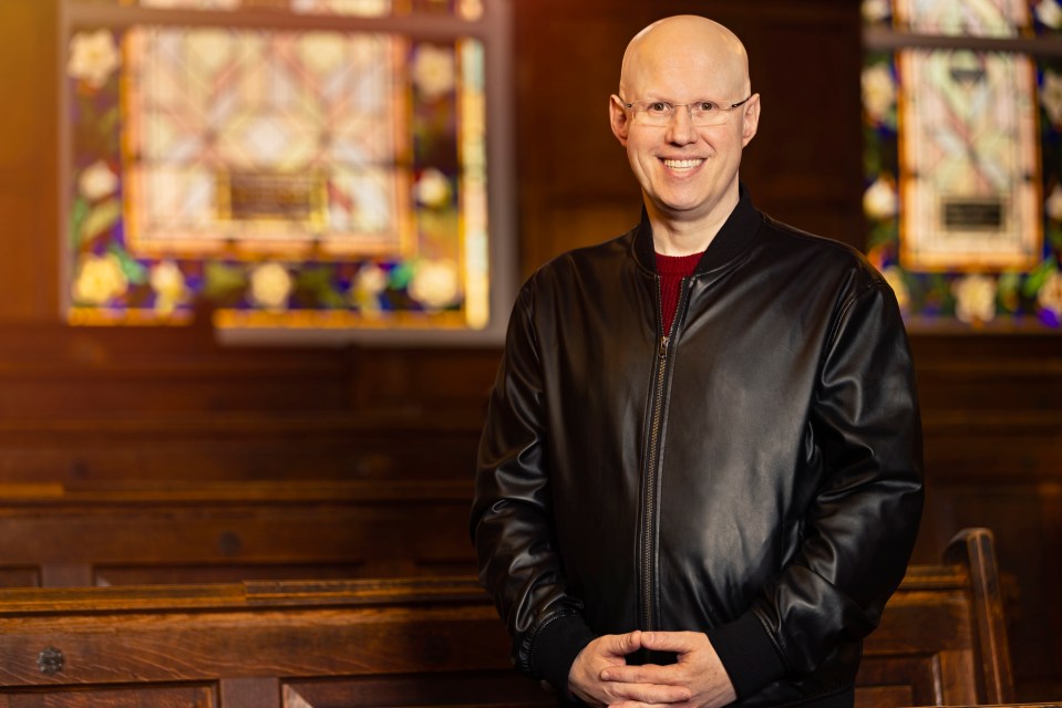 a man in a leather jacket stands in front of a stained glass window