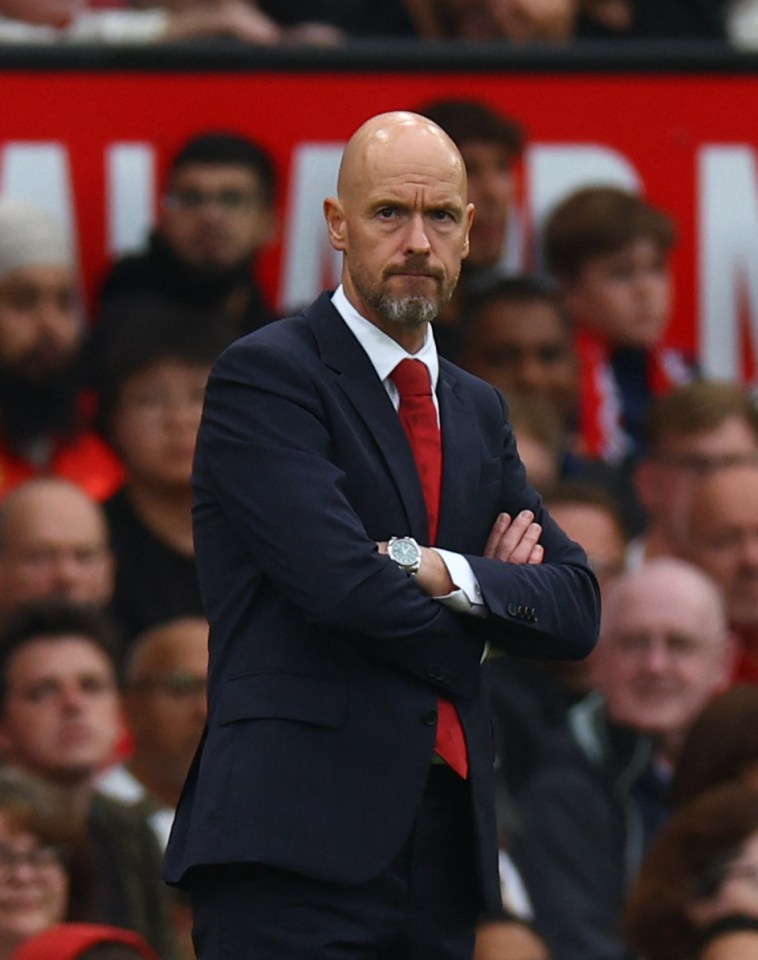 a bald man in a suit and red tie stands with his arms crossed