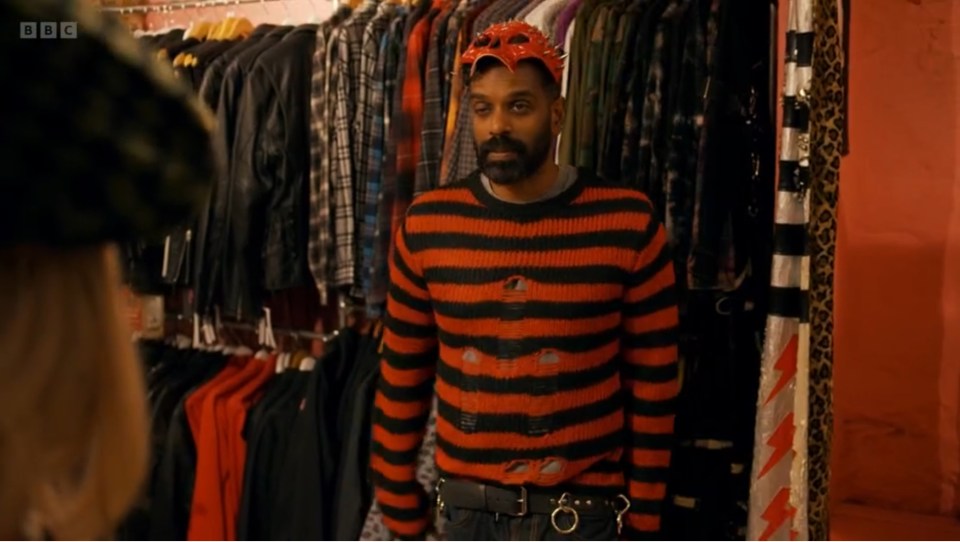 a man in an orange and black striped sweater stands in front of a bbc logo