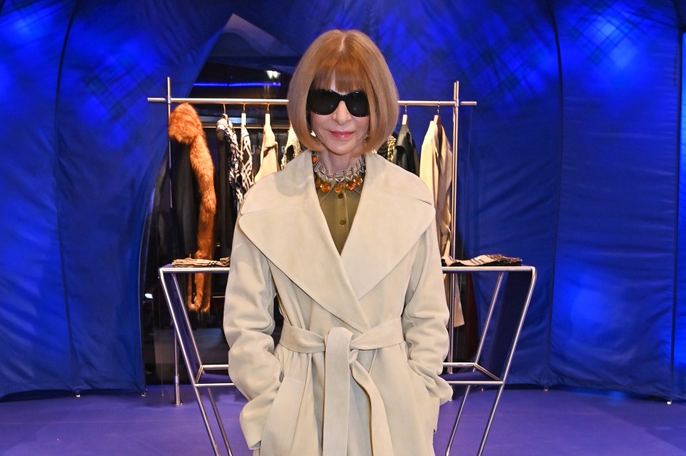 a woman wearing sunglasses and a white coat stands in front of a blue curtain
