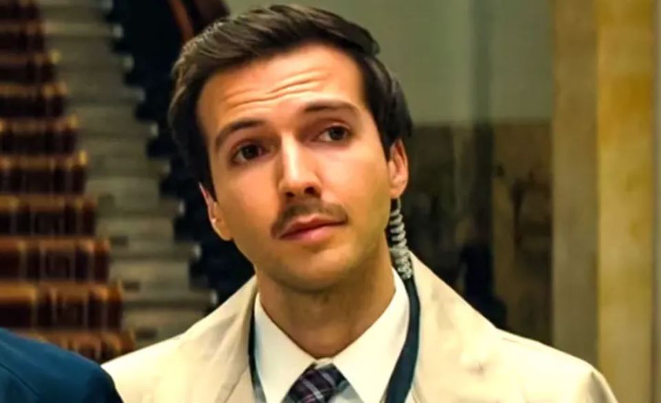 a man with a mustache is wearing a white coat and tie .