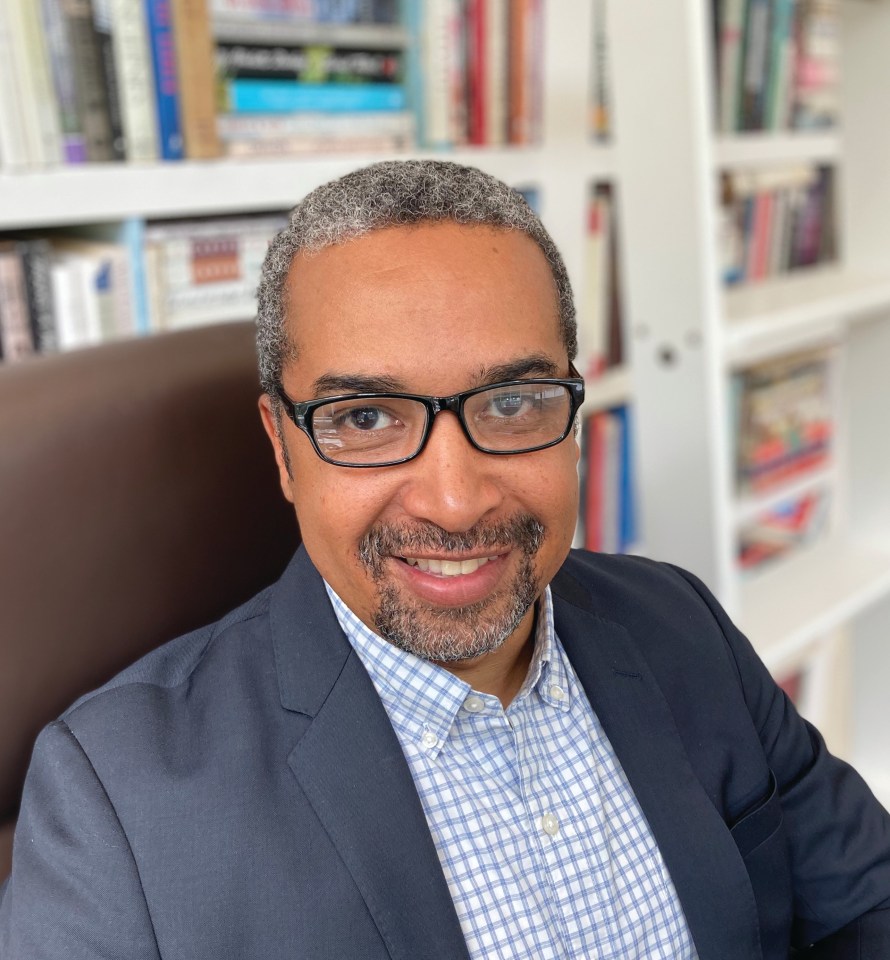 a man wearing glasses and a suit smiles in front of a bookshelf