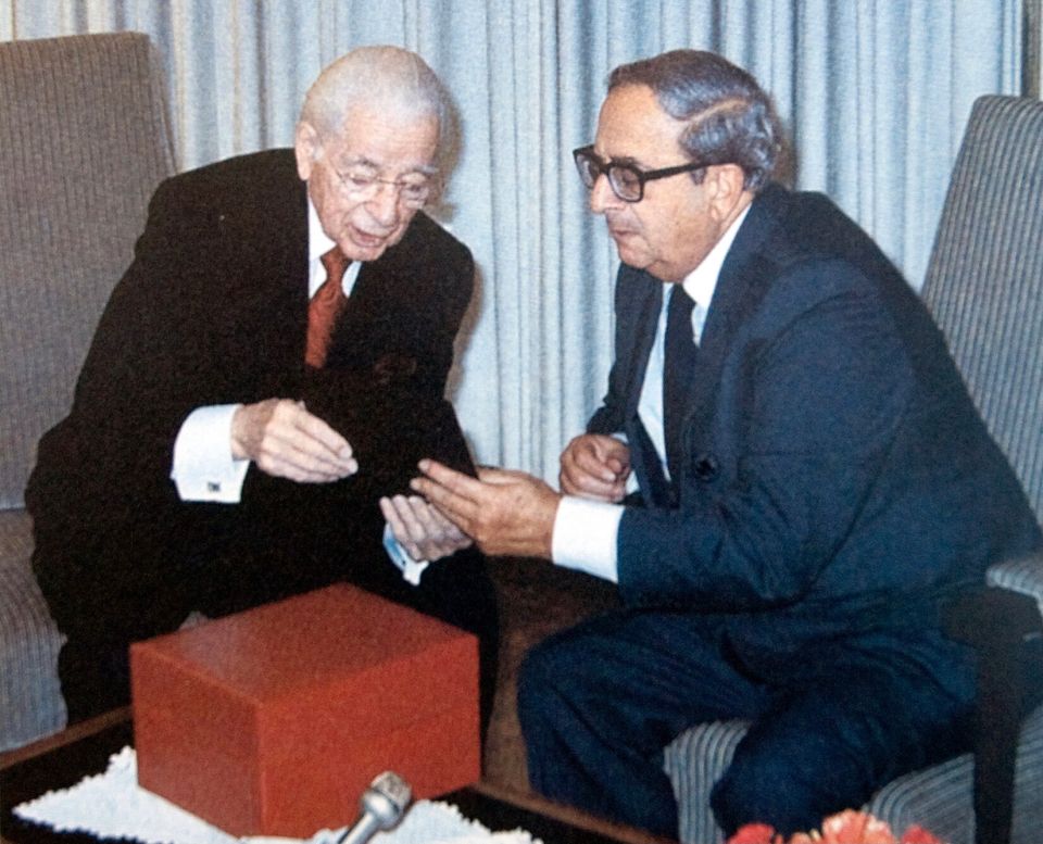 a man in a suit sits next to another man in a chair