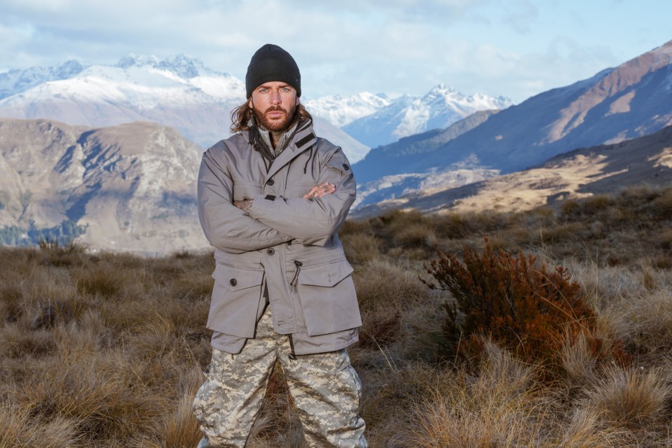a man with his arms crossed stands in a field with mountains in the background