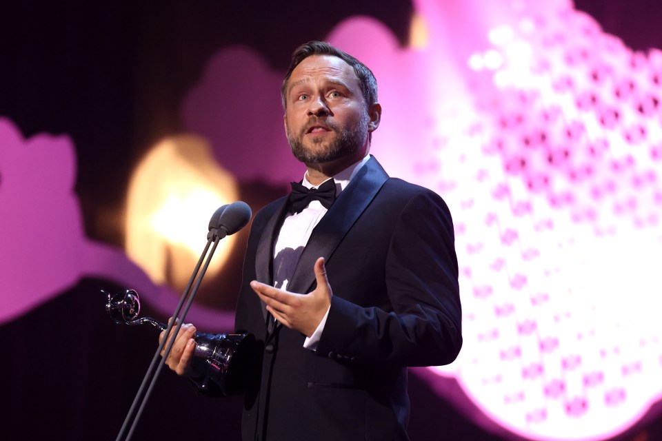 a man in a tuxedo holds a trophy in front of two microphones