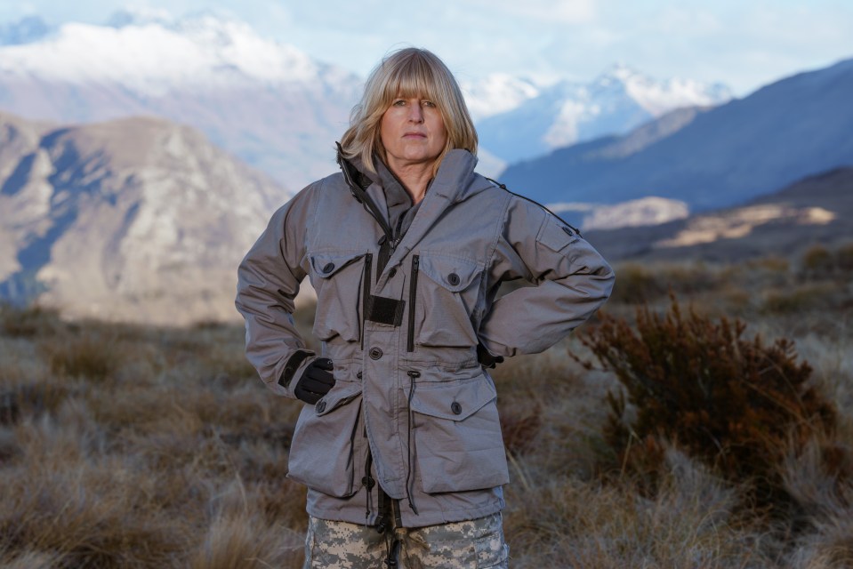 a woman in a camouflage jacket stands in a field with mountains in the background