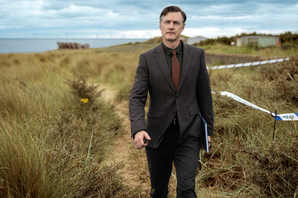 a man in a suit walks through a field with a police tape behind him