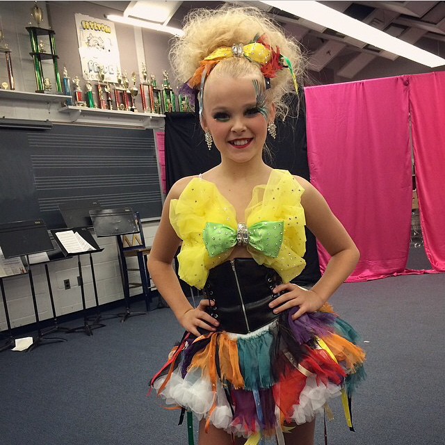 a girl in a colorful outfit stands in a room with trophies on the wall behind her