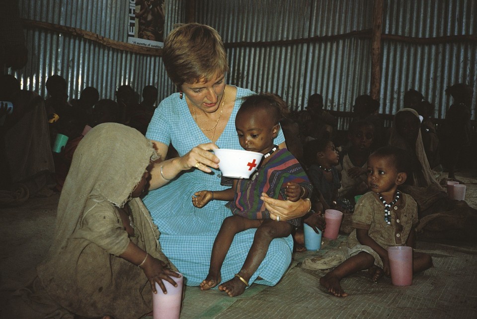 a woman is feeding a child from a bowl with a red cross on it