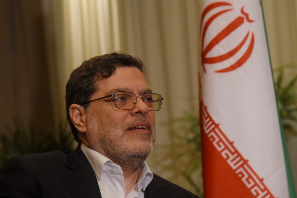 a man wearing glasses stands in front of an iranian flag