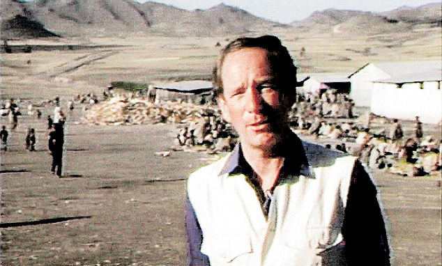 a man in a white vest stands in a field with mountains in the background