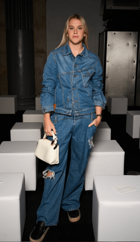 a woman wearing a denim jacket and jeans holds a white bag