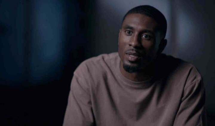 a man in a brown sweater is sitting in a dark room and looking at the camera .