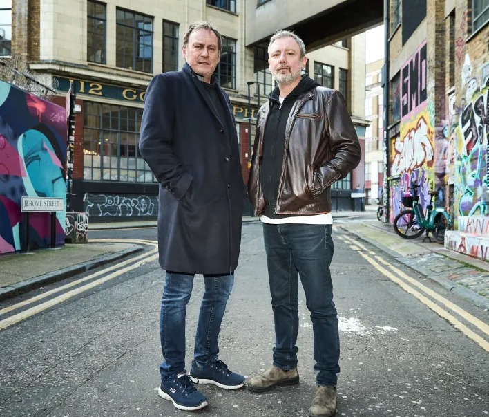 two men are standing on a street in front of a building with the number 12 on it