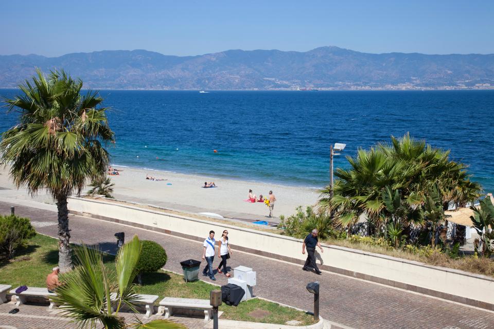 people walking on a sidewalk near a beach with mountains in the background