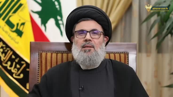 a man with a beard and glasses is sitting in a chair in front of a flag .