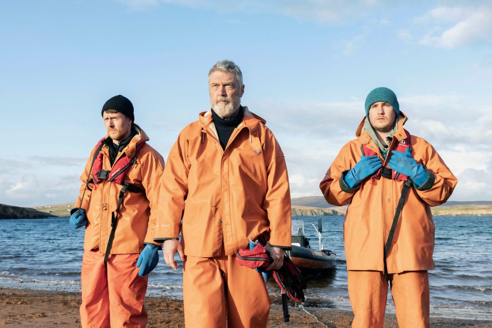 three men in orange jackets stand on a beach near a body of water