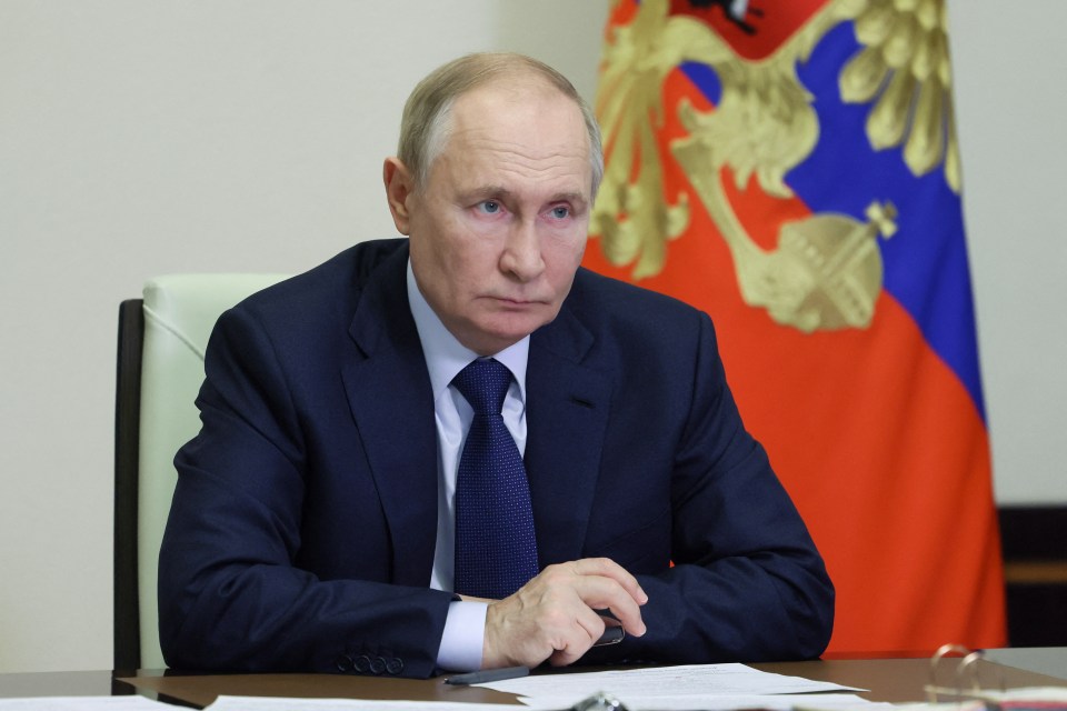 a man in a suit and tie sits at a desk in front of a russian flag