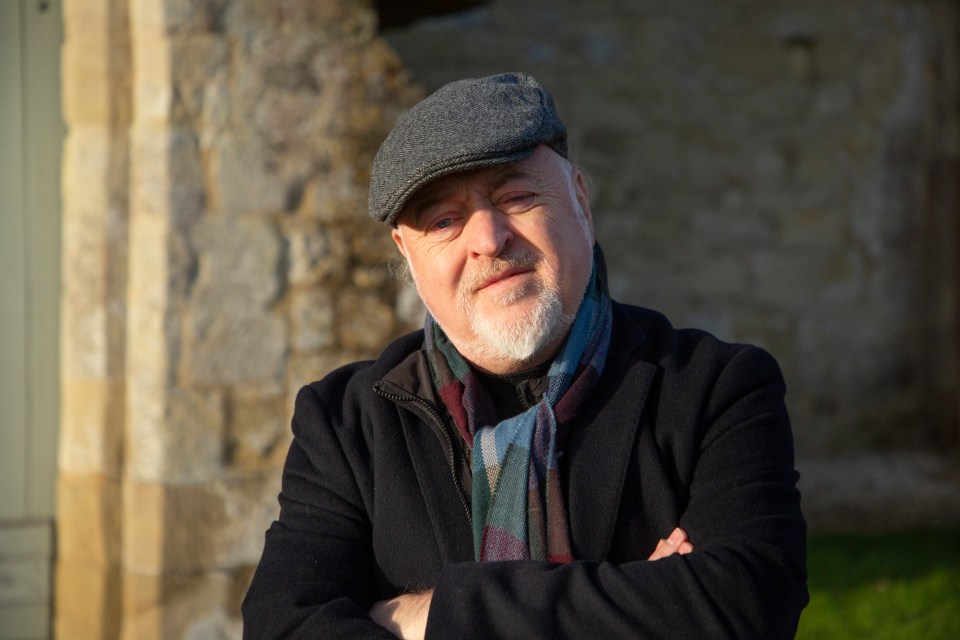 a man wearing a hat and scarf stands with his arms crossed