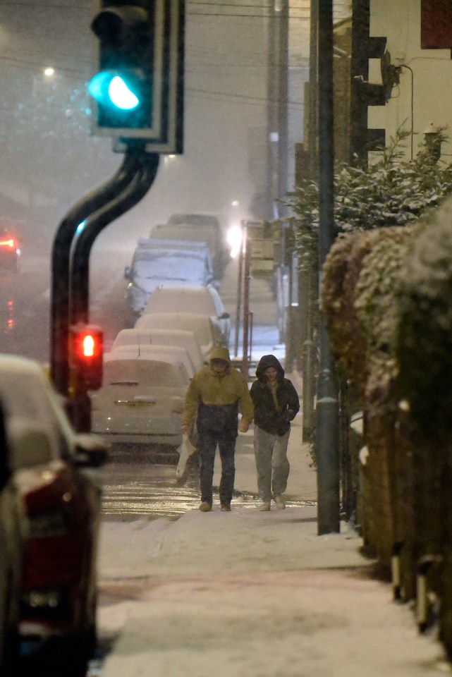 two people walking down a snowy street with cars covered in snow
