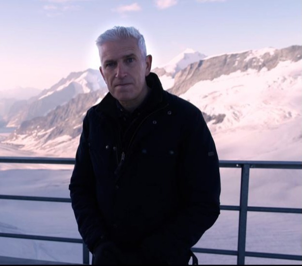 a man stands on a balcony overlooking a snowy mountain with the bbc logo in the background