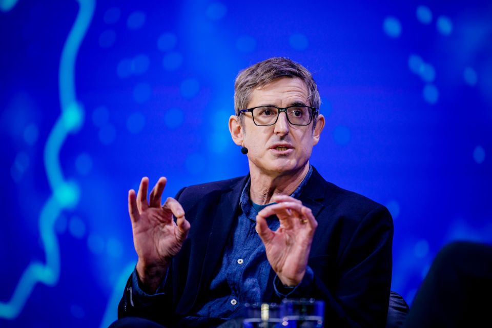 a man wearing glasses talks into a microphone in front of a blue background