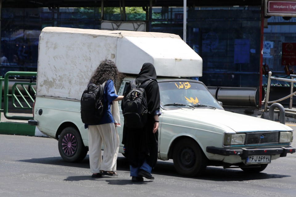 Two women, one wearing a hijab, walk past a pickup truck in Tehran, Iran.