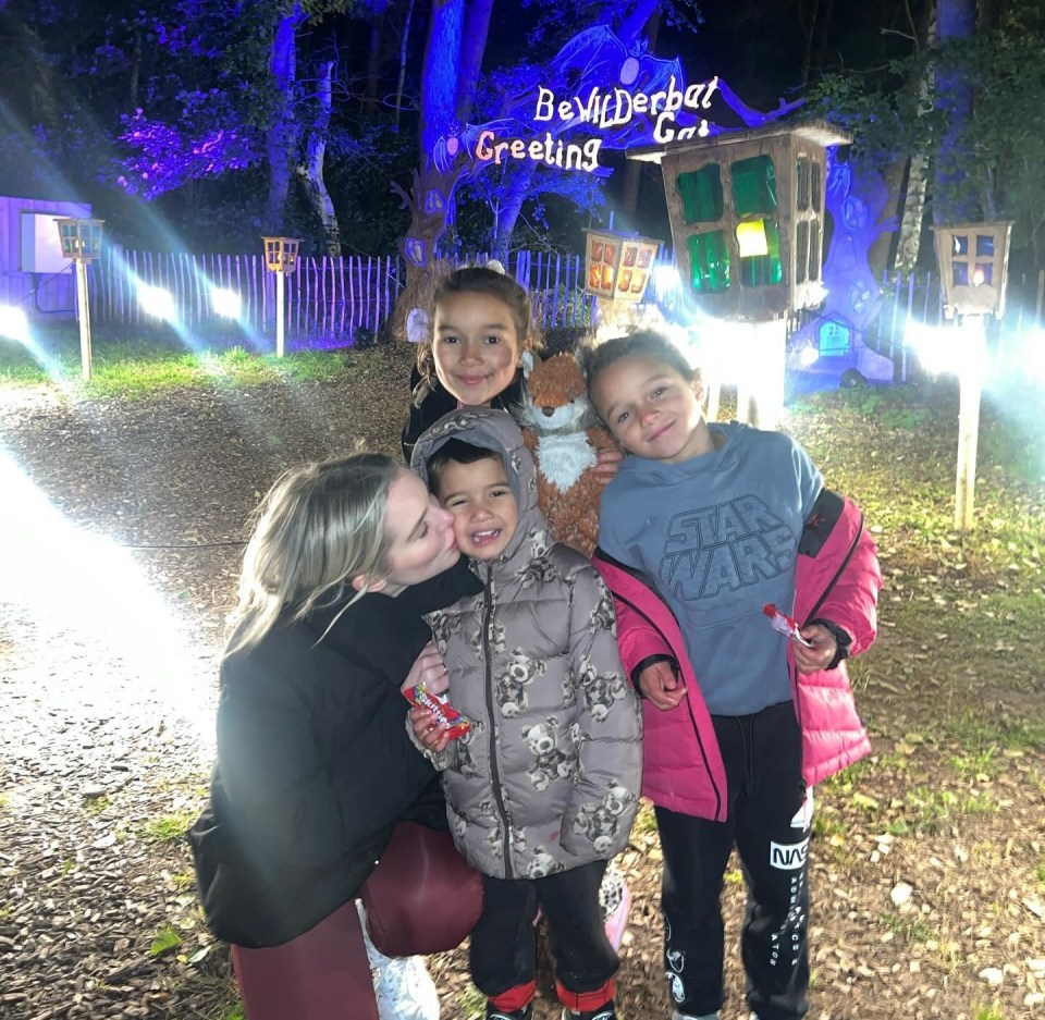 A woman kisses a young child while standing with two other children in front of a "Bewilderbat Greeting Gate" sign at night.